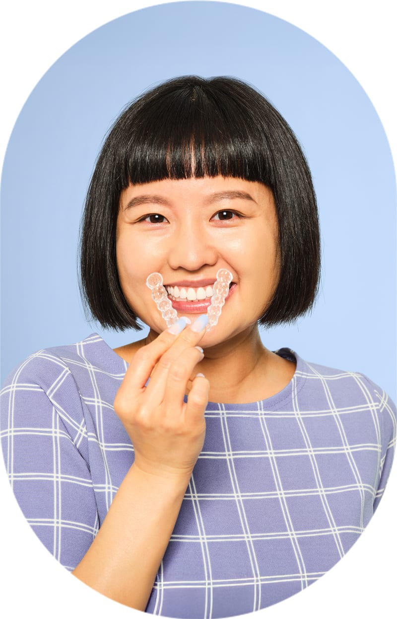A woman is smiling and holding a tray of clear aligners in front of her mouth.