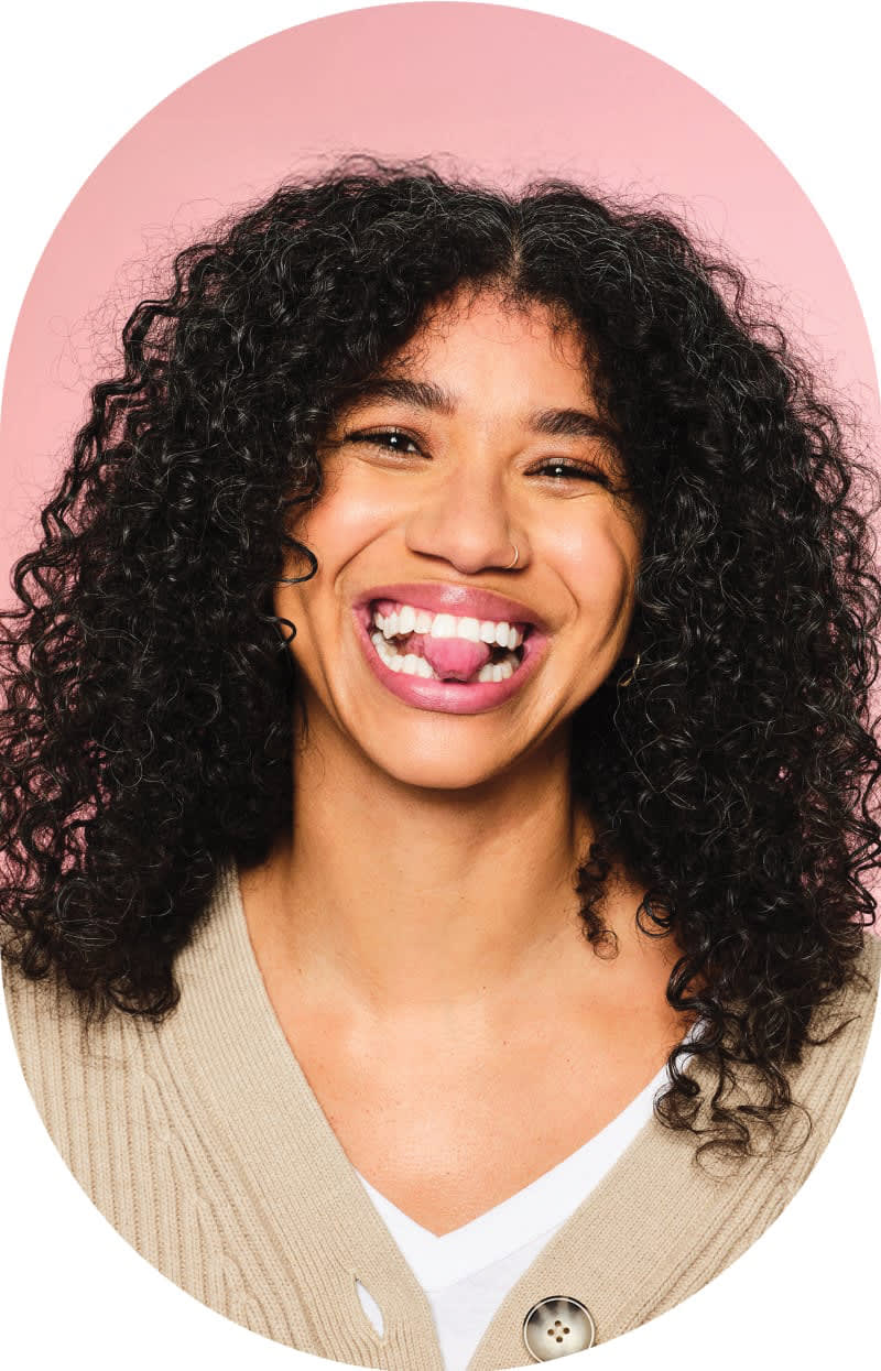 A woman with curly hair is smiling and biting her tongue.
