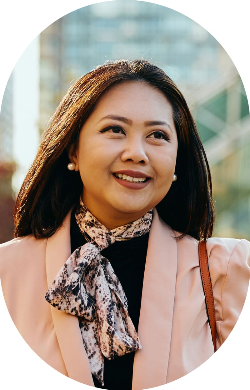 A woman in a pink jacket is smiling and looking to the right. She has a thin scarf around her neck.