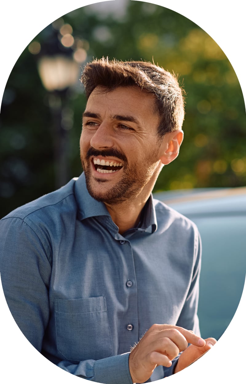 A man in a blue shirt is looking to the left and laughing. He is leaning against a car.