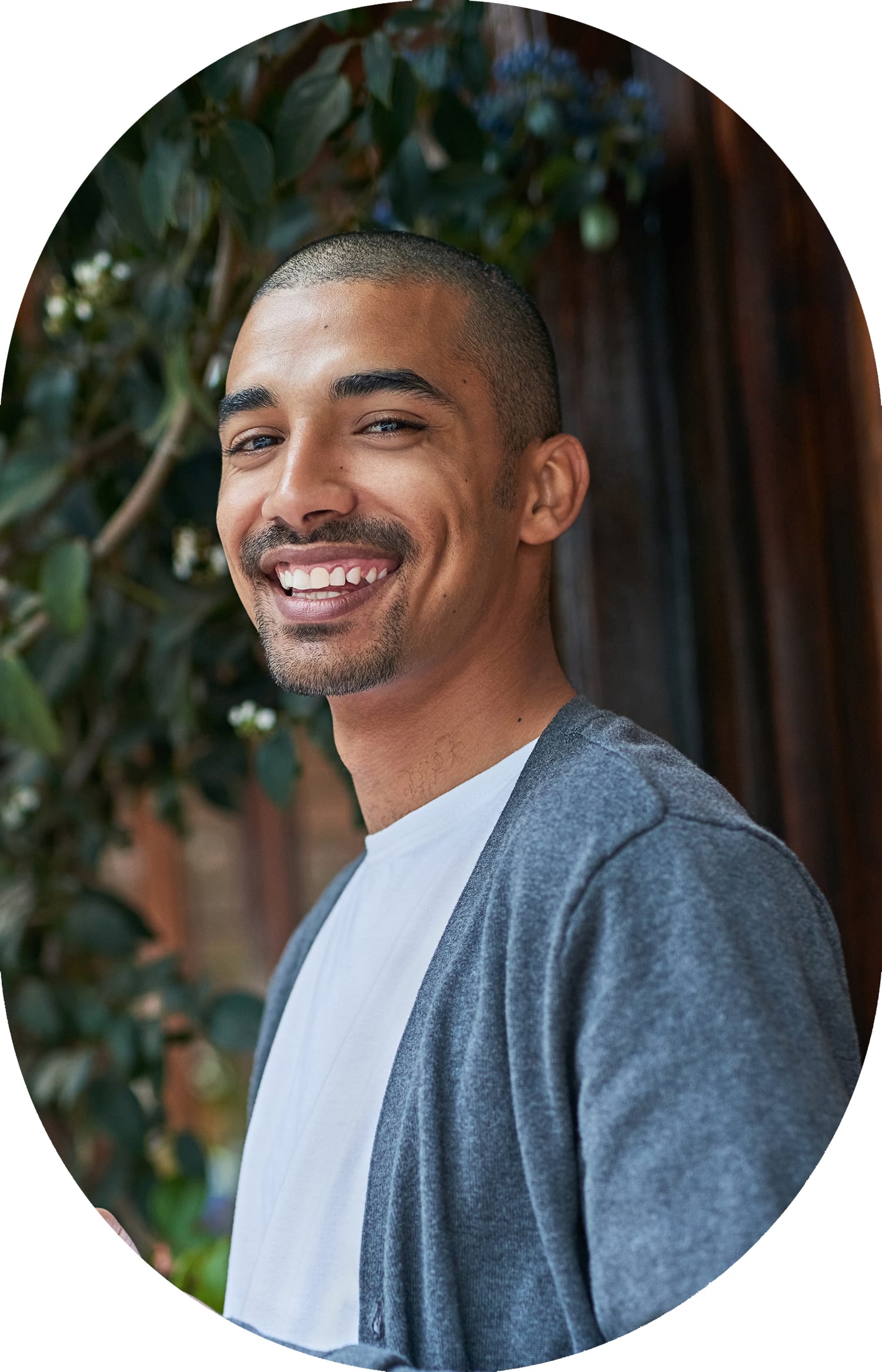 A smiling man stands in front of a wooden fence, with a tree to his right.