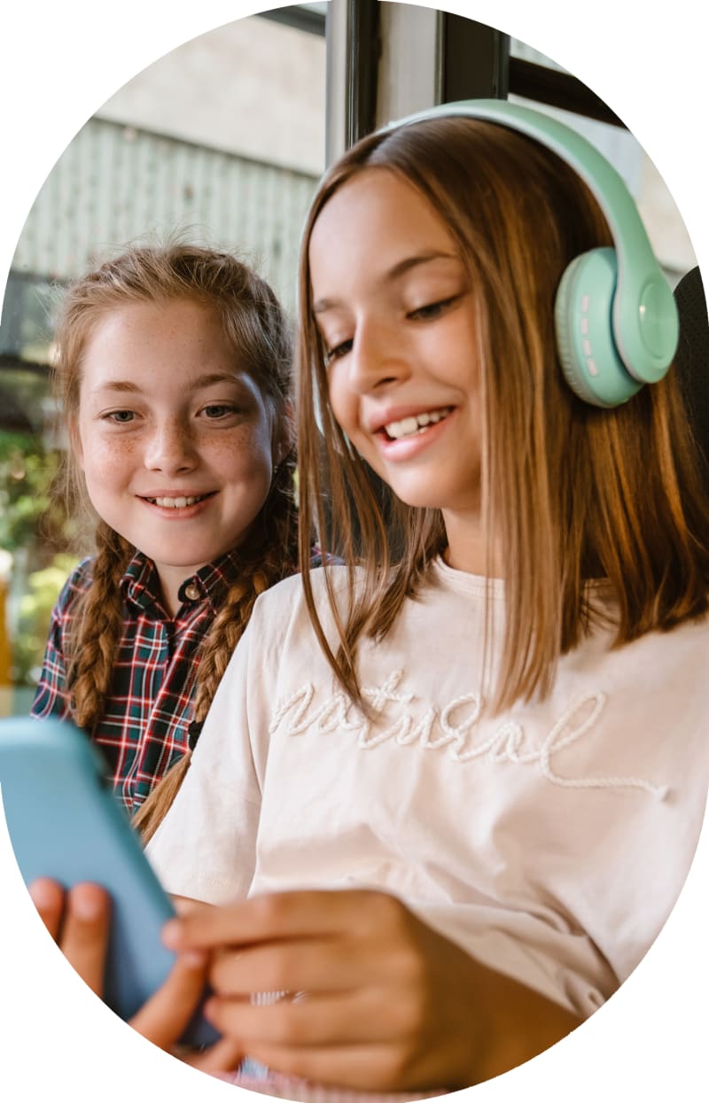 Two young girls are sitting together on a school bus, laughing. One is wearing headphones.