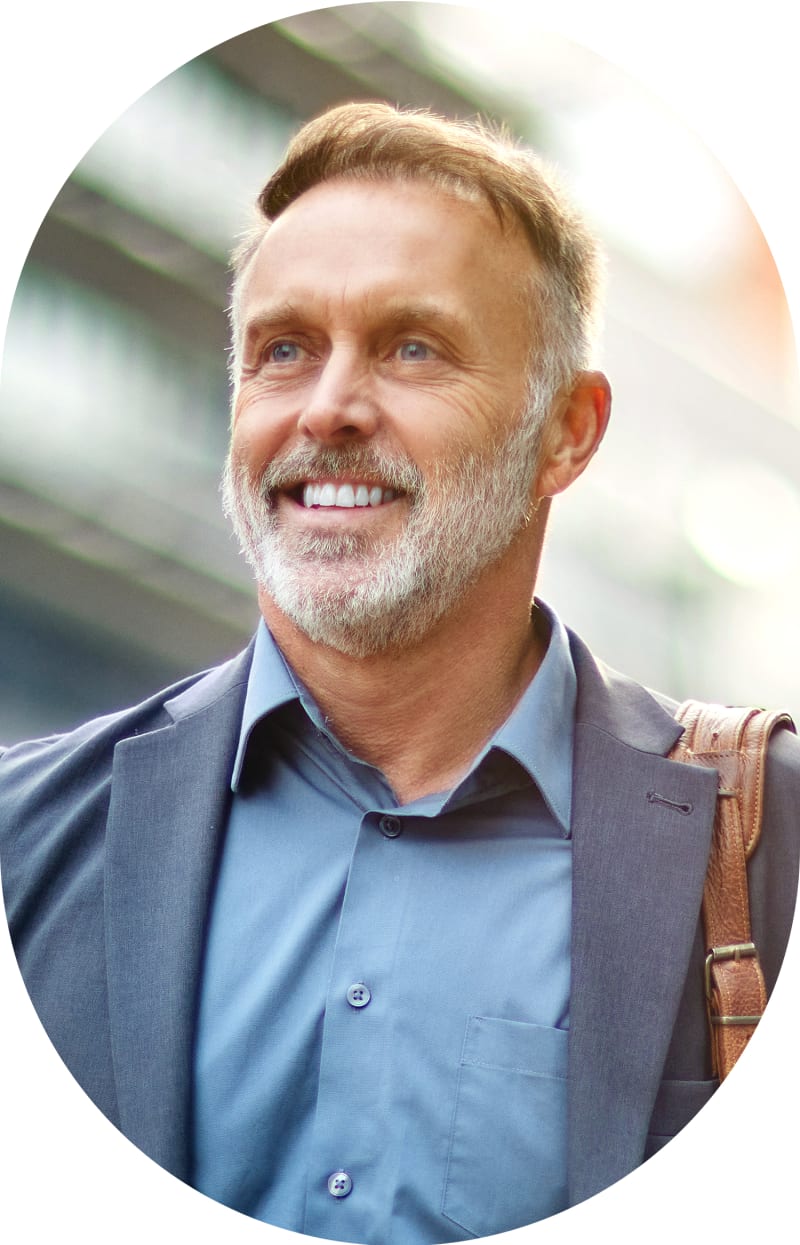 A bunsiness man with a bag strap over his shoulder stands outside smiling.