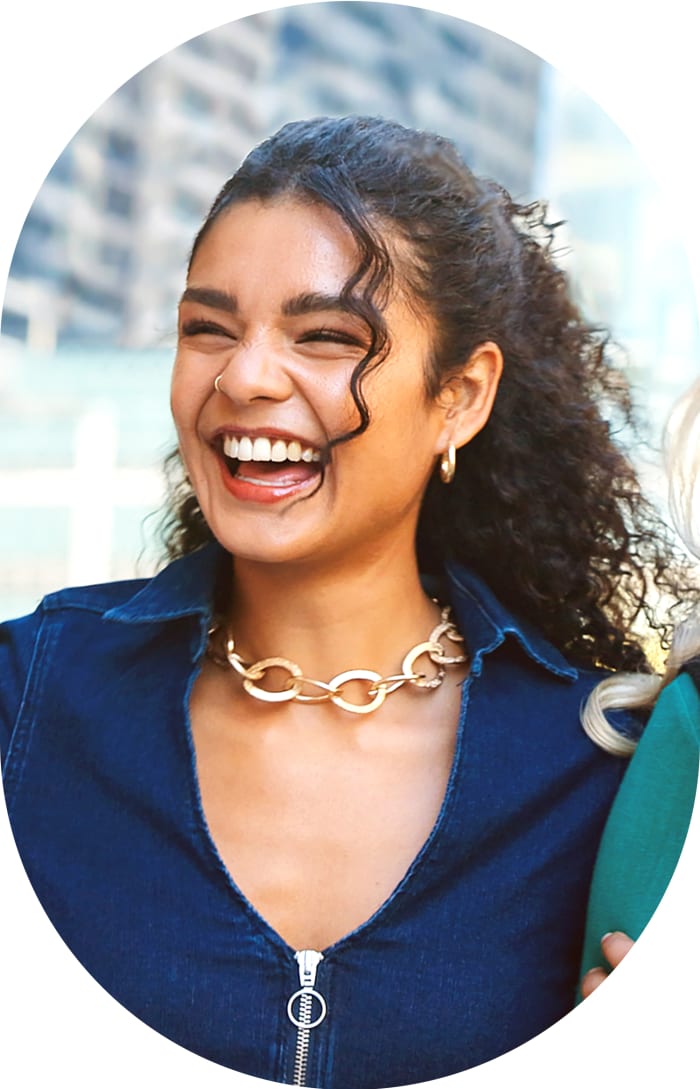 A young woman in a blue zip-up top is standing outside laughing and smiling.