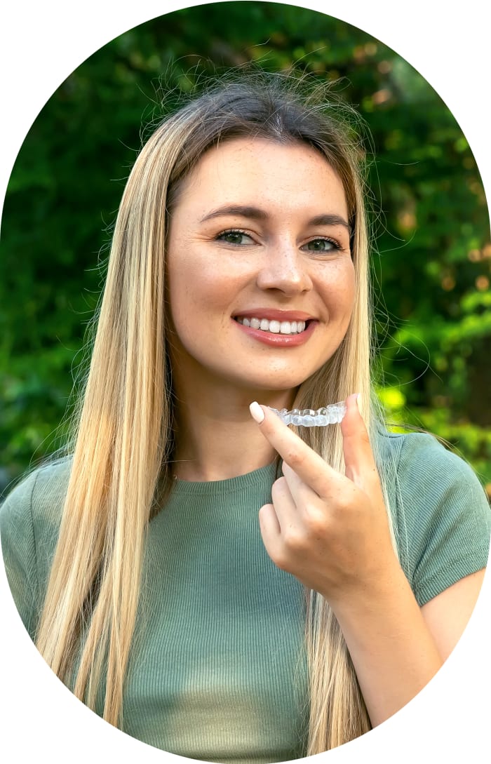 A woman stands outside smiling while holding up a clear aligner.