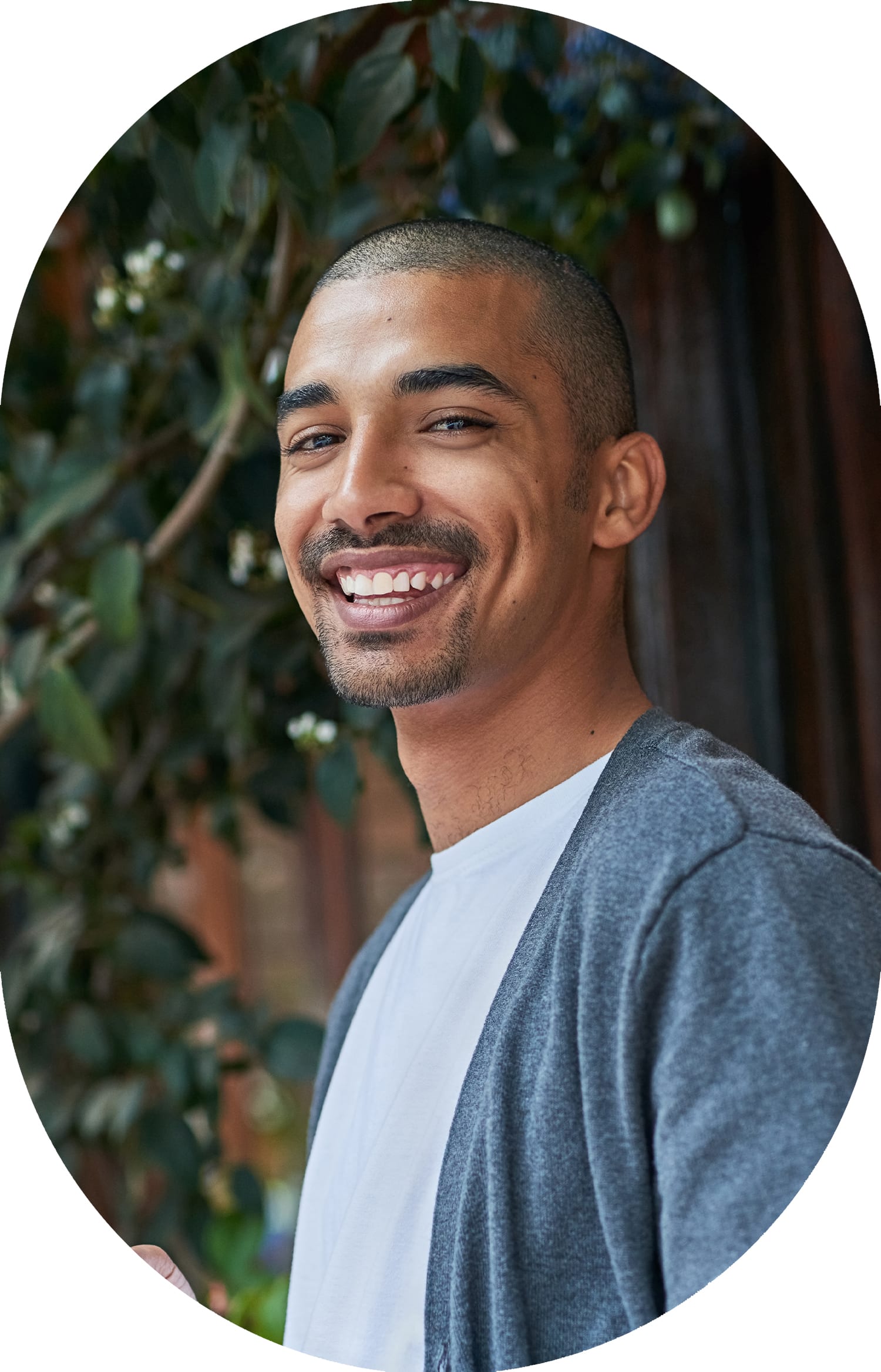 A young man in a grey shirt with a darker grey cardigan over it is standing in front of some foliage and smiling down at the camera.