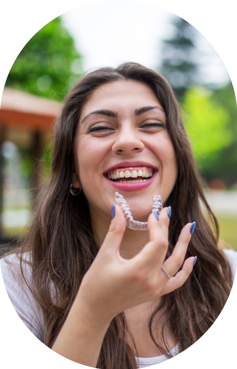 A young woman is smiling and holding a clear aligner tray in front of her mouth.