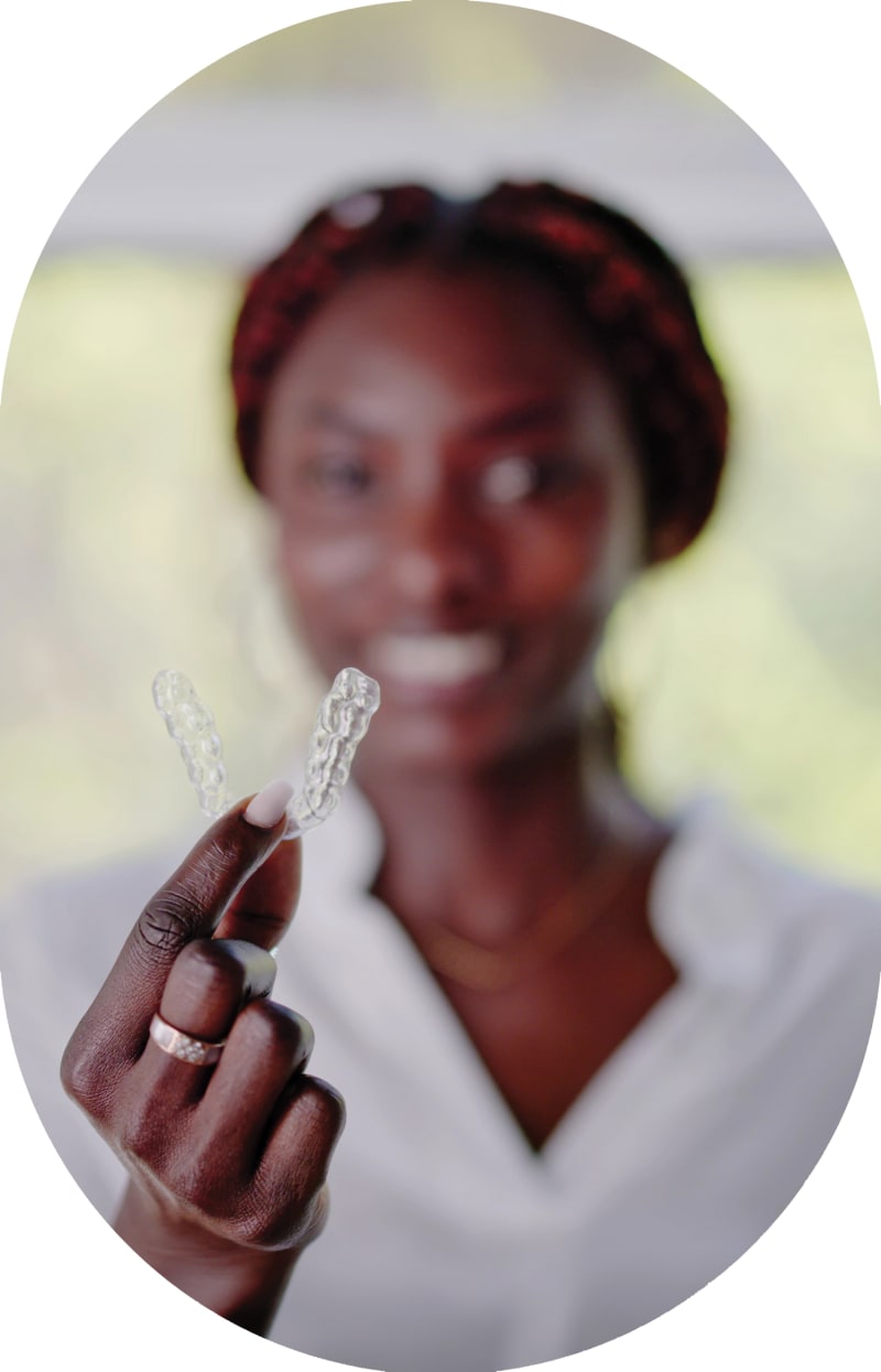 A woman is standing out of focus in the background. In the foreground, she holds a clear aligner tray.