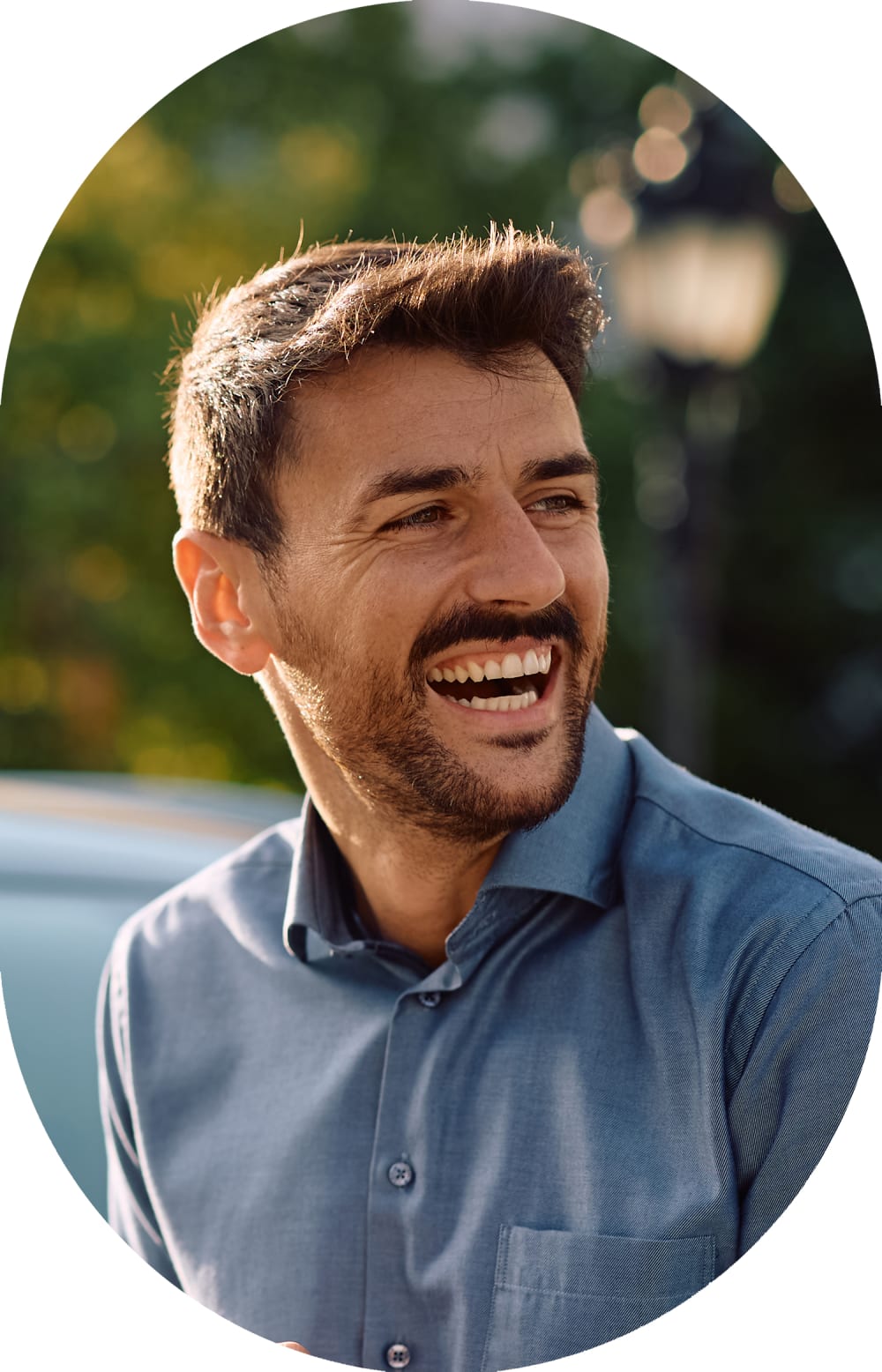 A bunsiness man with a bag strap over his shoulder stands outside smiling.