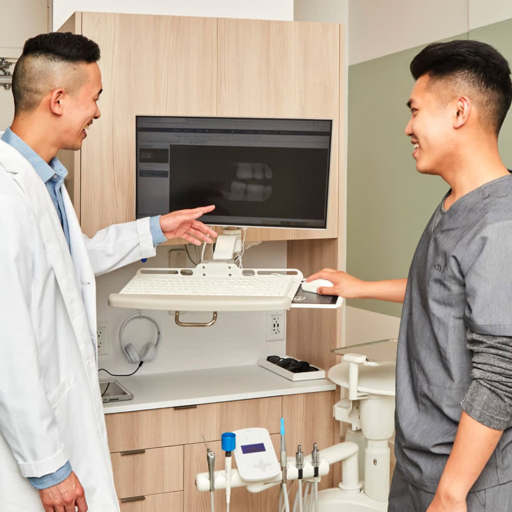 Male dentist pointing to a model of teeth while explaining treatment to a patient seated in the dental chair.