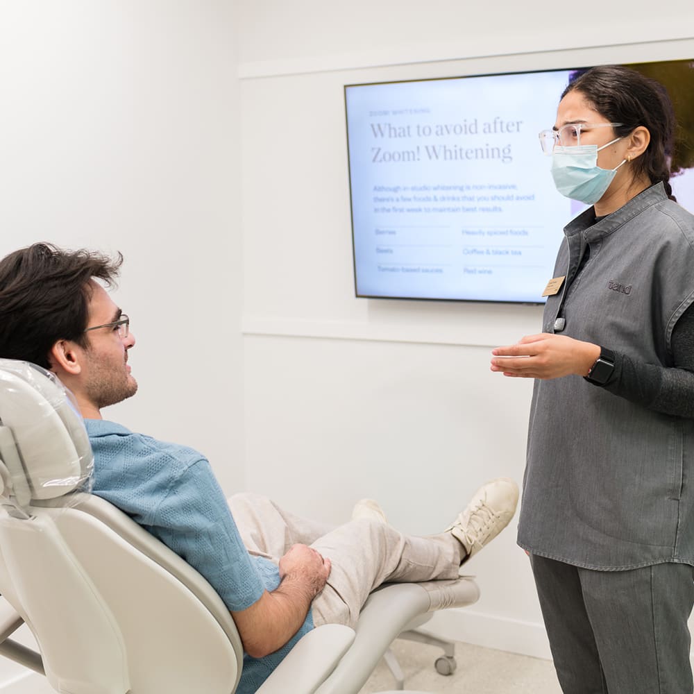 Male dentist pointing to a model of teeth while explaining treatment to a patient seated in the dental chair.