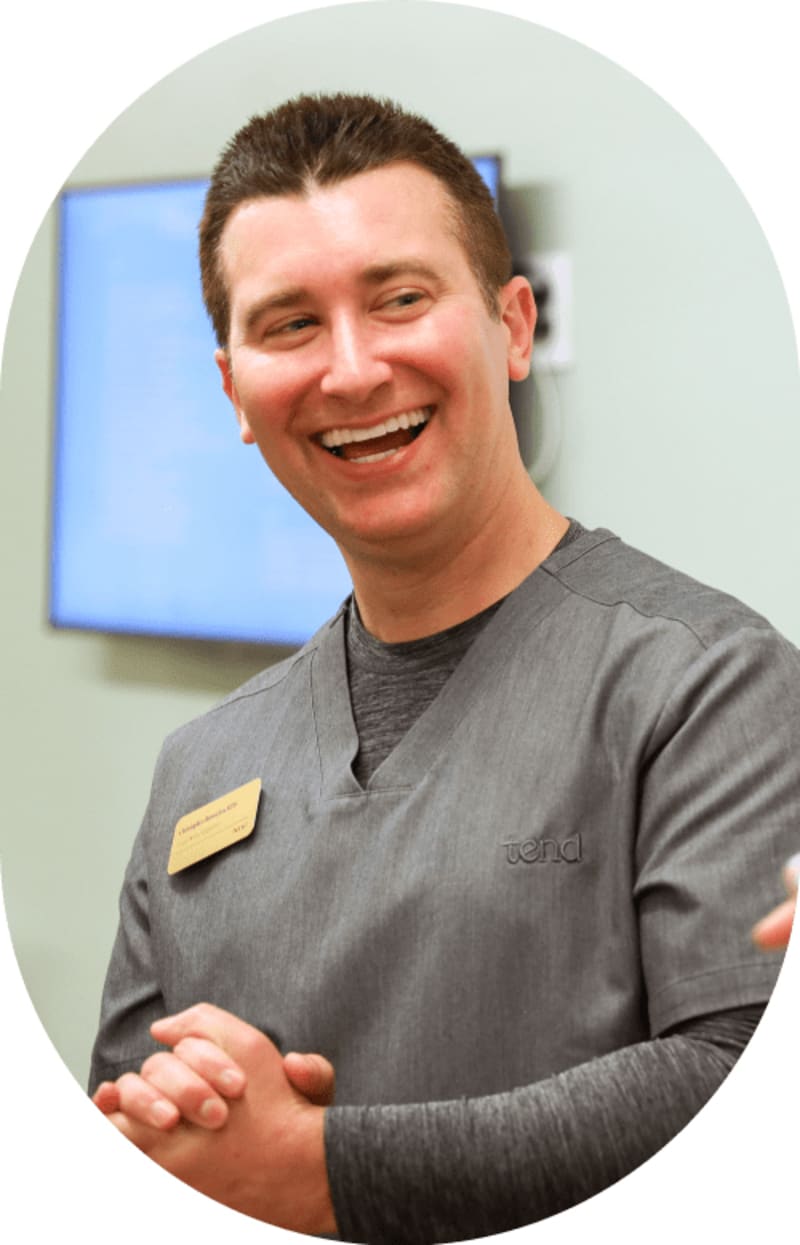 Smiling male team member with hands clasped, standing in front of a treatment room screen, looking to his left.