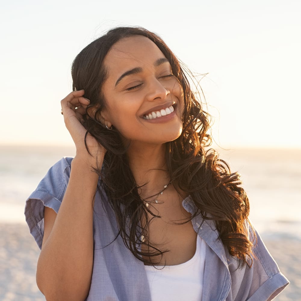 A woman with long hair stands outside smiling with her eyes closed and tucking hair behind her ear.