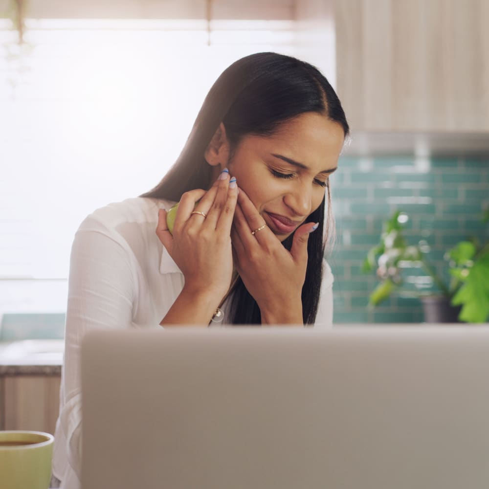 A woman is sitting at her laptop. She is holding her cheek, indicating tooth pain.