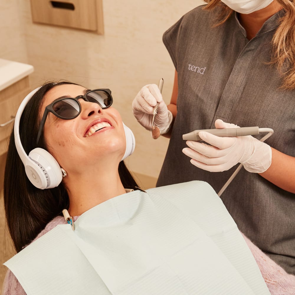 A female patient is sitting in an exam chair with headphones and sunglasses on. A dental professional is sitting beside her with tools in her hands, ready to start the exam.