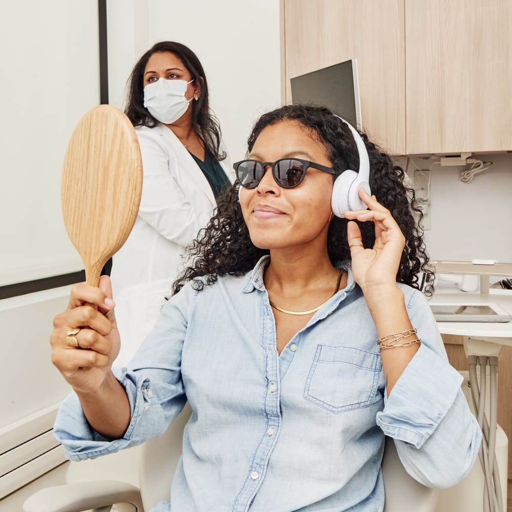 A woman sits in the dental treatment chair looking at herself in a handheld mirror while the dentist stands behind her.