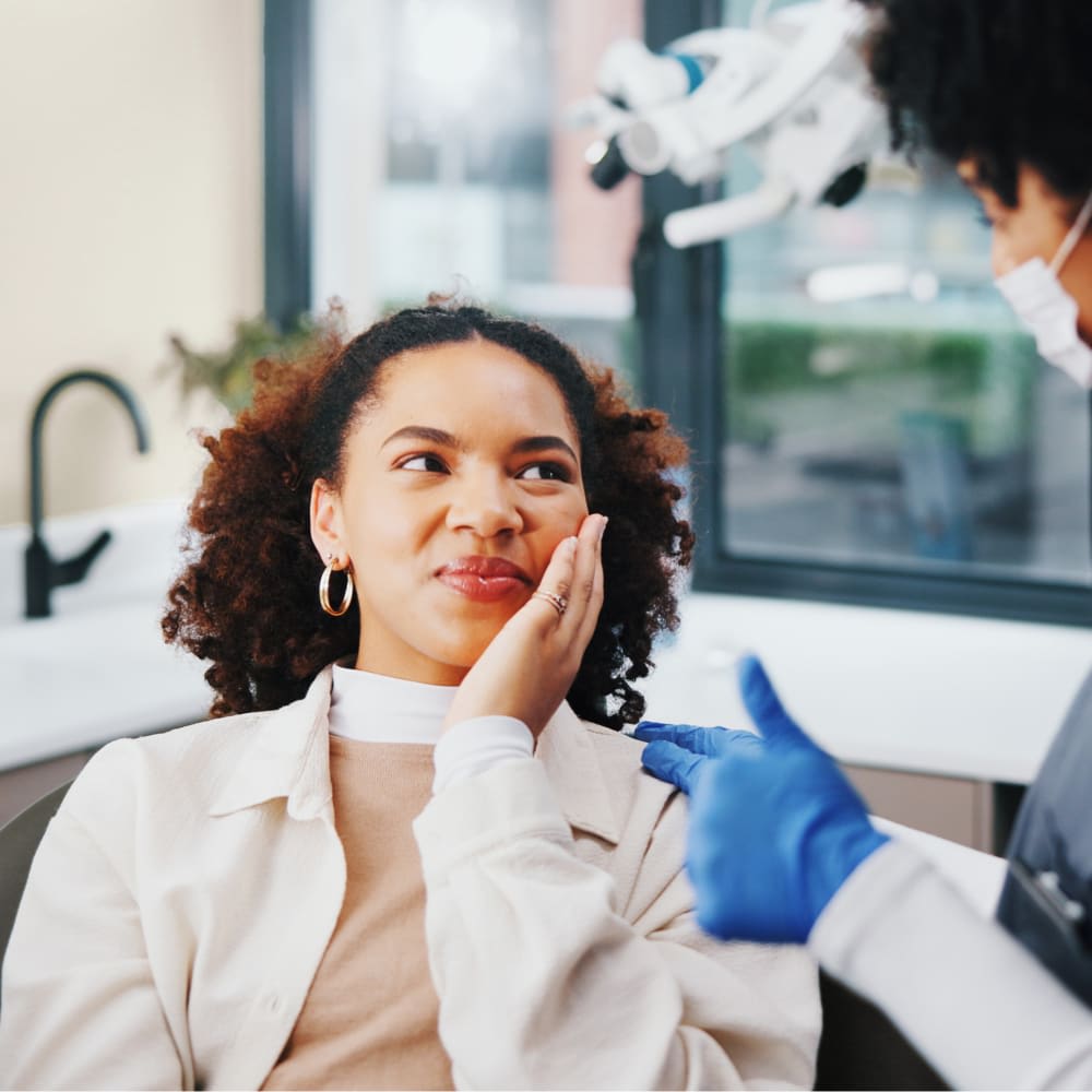 A woman sits in a dental treatment chair, lookign the dentist and holding the side of her face.