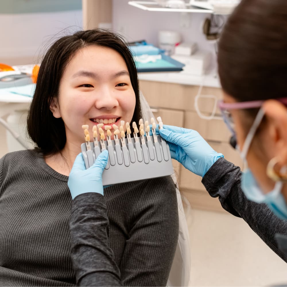 A dental professional is holding up a model of different shades of white to a female patient's teeth to determine which shade will suit her best.