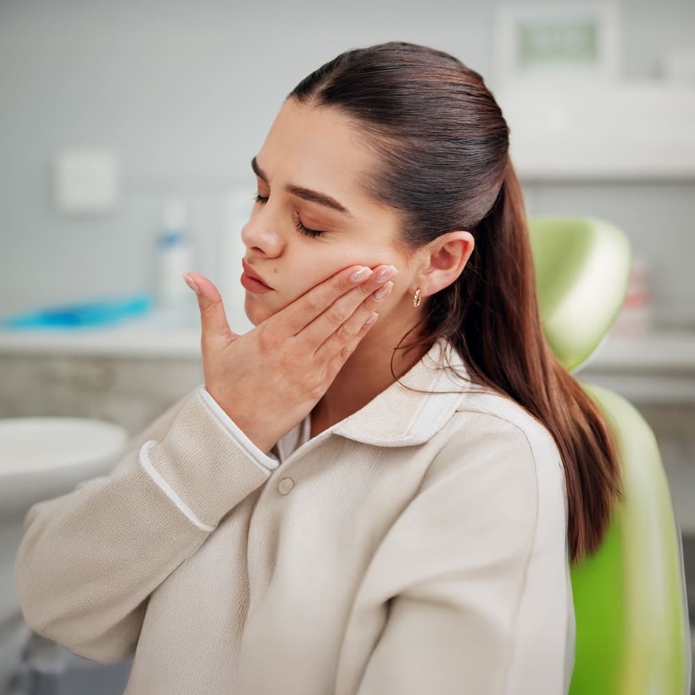 A young woman is sitting in a dental chair, holding her face like her tooth hurts.